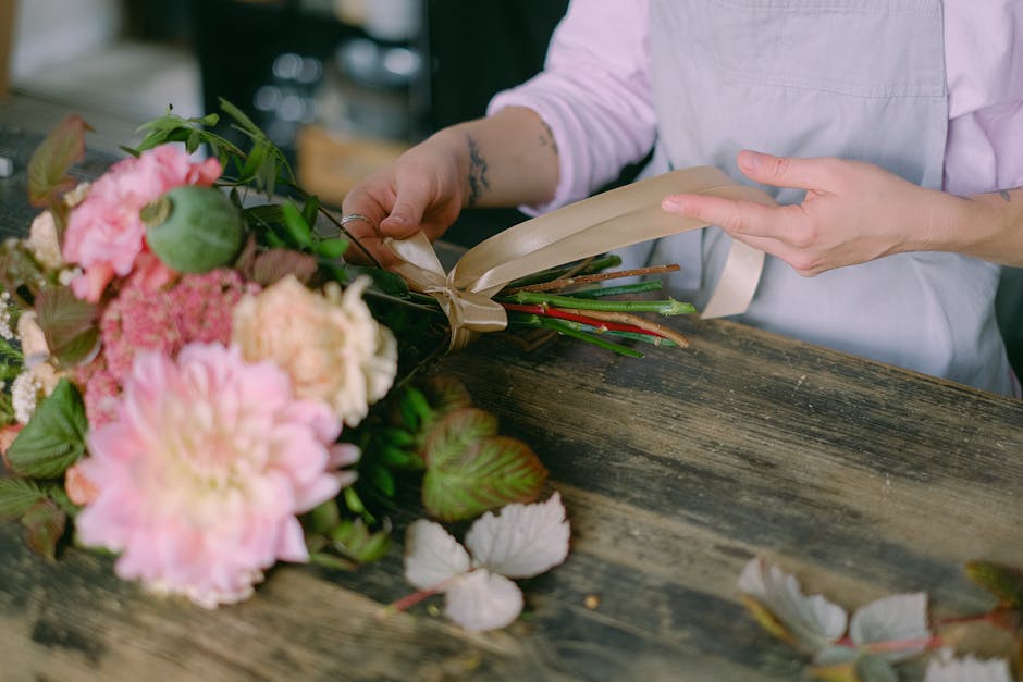 A florist wearing a light pink shirt and an apron is seen preparing a floral bouquet arrangement on a rustic wooden table. The bouquet features large, soft pink peonies and clusters of pink and cream 
