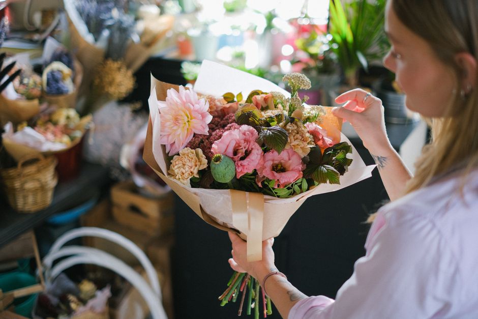 A woman with long blonde hair, wearing a light-colored shirt, is holding a large bouquet of fresh flowers wrapped in beige paper and tied with a beige ribbon. The bouquet includes soft pink dahlias, p