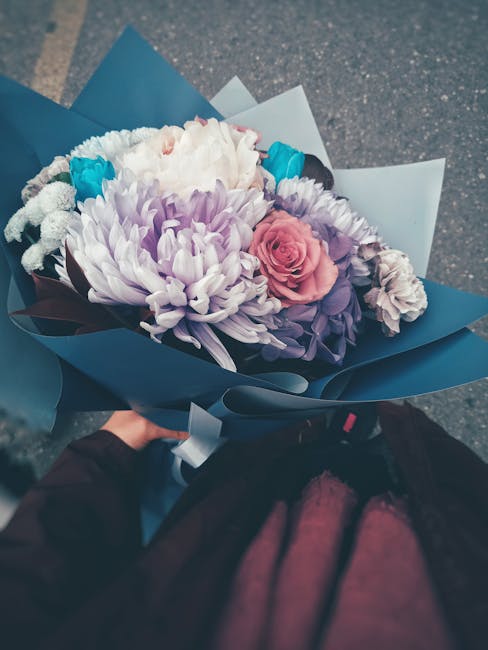 A hand holding a floral bouquet arranged with a variety of fresh flowers, including large white and lavender chrysanthemums, a pink rose, and smaller blue and purple flowers, wrapped in elegant blue a