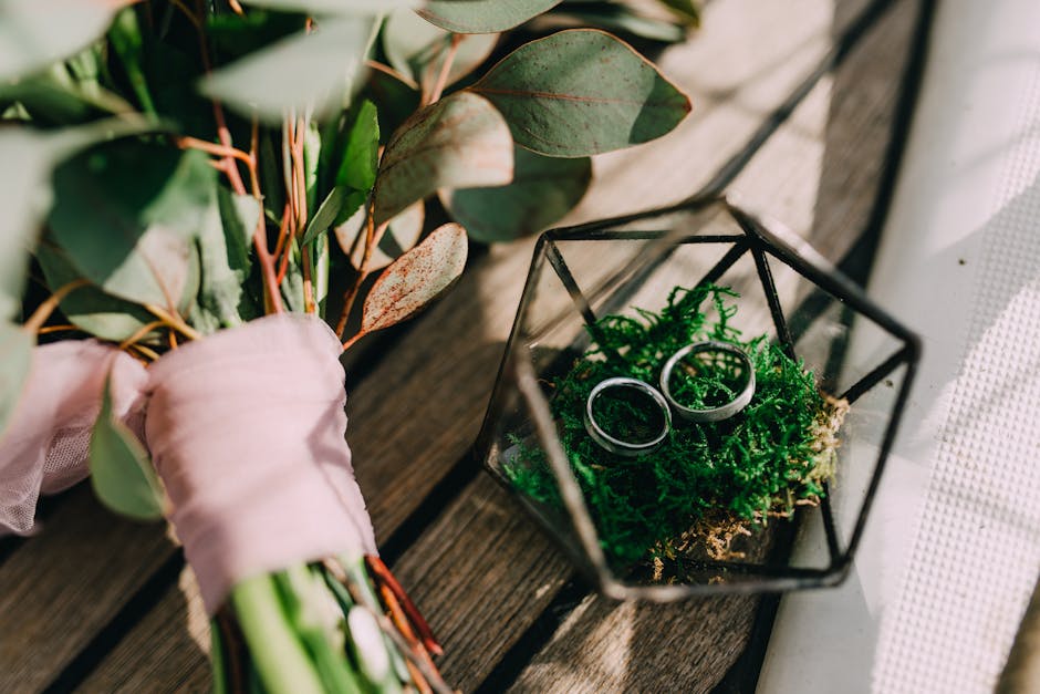 A close-up of a bridal bouquet resting on a rustic wooden surface, featuring an arrangement of eucalyptus leaves with green and reddish-brown tones, wrapped with soft pink ribbon. Adjacent to the bouq