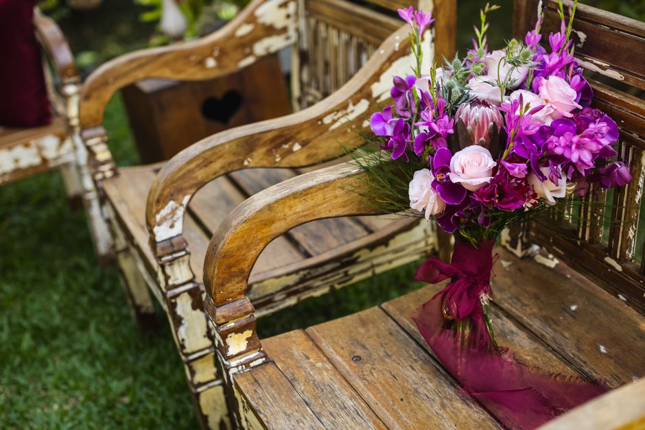 A bouquet arrangement featuring pink and purple flowers, including roses, orchids, and hydrangeas, is placed on an antique wooden garden bench with chipped white paint, secured with a deep pink ribbon